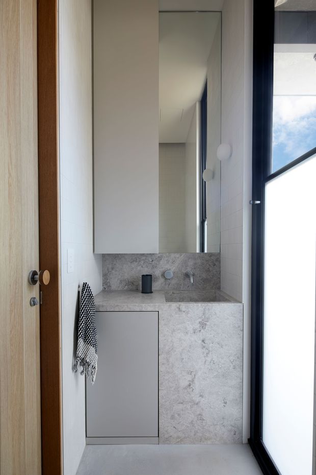 Compact contemporary bathroom vanity viewed from the doorway. The space features a wall-mounted grey vanity with a pale grey stone-look benchtop and matching side panel, integrated basin and minimalist chrome tapware. A large frameless mirror extends above the vanity, while white tiled walls, a pale floor, timber door, and black-framed full-height window create a bright, restrained interior.