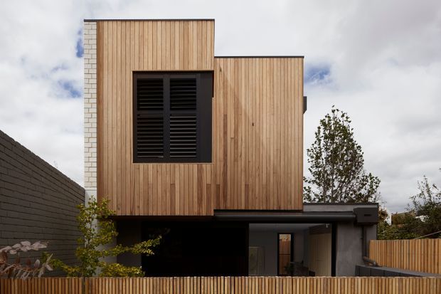 Exterior view of a compact contemporary multi-level home with a strongly articulated box-like form. The upper storey is clad in vertical warm timber boards, with a contrasting dark louvred window and a projecting lower-level entry or carport zone. A pale masonry side wall and timber boundary fencing frame the house.