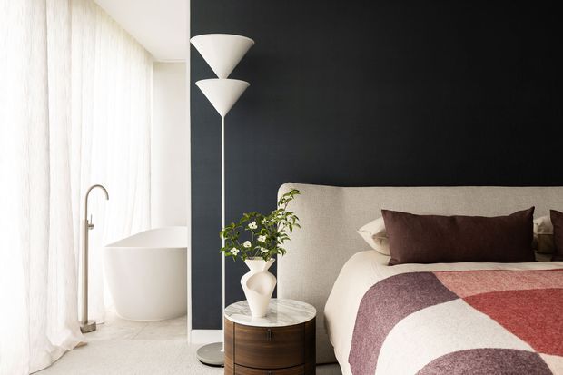 A refined contemporary bedroom with a partially visible freestanding white bathtub beside sheer floor-to-ceiling curtains. The bedroom features a dark charcoal accent wall, upholstered cream bedhead, layered cream and burgundy bedding, a sculptural white floor lamp, and a round timber bedside table with flowers and decor.