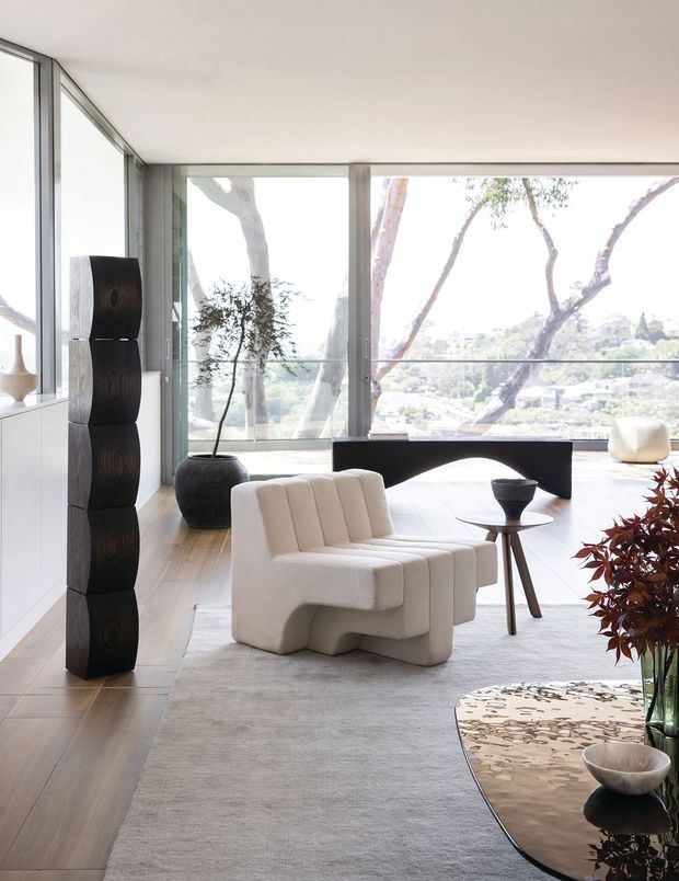 A bright, refined contemporary living room with expansive floor-to-ceiling glazing overlooking a leafy suburban landscape. The room features pale timber flooring, white built-in cabinetry, a sculptural cream upholstered lounge chair, a dark sculptural console, a small side table, neutral carpeting, and carefully curated dark and natural-toned decorative objects. Large windows and an open arrangement create an airy, gallery-like atmosphere.
