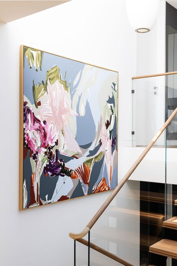 Bright modern stairwell featuring a large colourful abstract floral painting in a timber frame, white walls, glass balustrades, and a slim timber handrail with black metal supports. Timber stair treads and a contemporary pendant light are visible, creating a clean, airy interior.