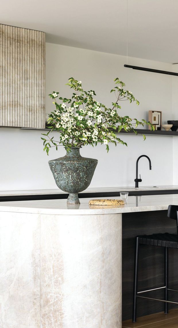 A refined contemporary kitchen featuring a large curved island clad in pale veined stone, a dark timber or black base, and a black gooseneck tap. A sculptural mottled ceramic vase with flowering branches forms the centrepiece on the island. Behind it are dark open shelves, pale cabinetry with vertically fluted detailing, and a restrained neutral palette. A black bar stool is partially visible at the island.