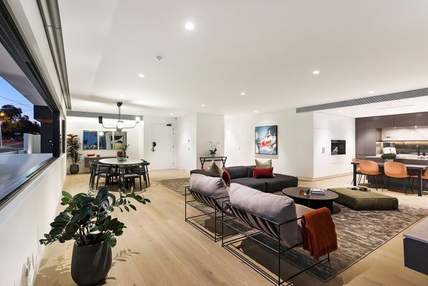 Open-plan contemporary apartment interior combining a living room, dining area and kitchen. The bright space has white walls and ceiling, pale timber flooring, large windows, recessed lighting, a black-framed lounge suite, dining table, and a modern kitchen with dark cabinetry and a light benchtop.