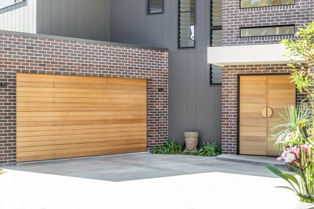 Contemporary residential facade featuring dark grey vertical cladding, red-brown brickwork with light mortar, warm horizontal timber garage and entry doors, and a broad concrete driveway. Narrow horizontal windows and simple geometric detailing reinforce the modern exterior, with limited planting at the entry.