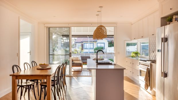 Bright open-plan kitchen and dining area with white shaker-style cabinetry, a central island, stainless-steel appliances, timber flooring, and a timber dining table. Large glazed doors connect the room to an outdoor or adjoining living area, while a woven pendant light adds warmth.