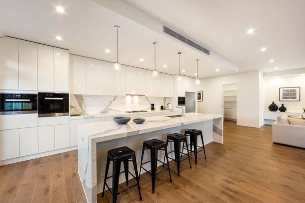 Bright open-plan contemporary kitchen with a large white marble-look island, four black bar stools, glossy white floor-to-ceiling cabinetry, integrated appliances, pendant lighting, and warm timber flooring. A living area and hallway are visible beyond the kitchen.