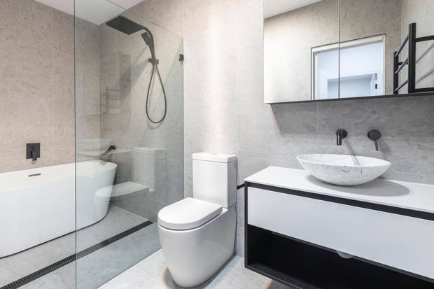 Contemporary bathroom with a freestanding bathtub behind a frameless glass shower enclosure, wall-mounted rainfall shower, sculptural white toilet, and floating vanity with a vessel basin. Large-format light grey textured wall and floor surfaces create a clean, refined monochromatic scheme, with black tapware, shower fittings, shelving, and trim providing contrast.