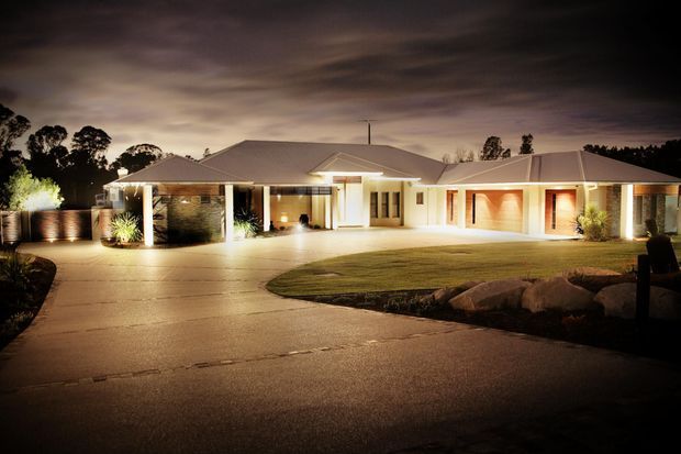 A single-storey suburban residence photographed at dusk, with a broad low-pitched tiled roof, multiple gabled roof sections, pale rendered walls, dark stone feature walls, and warm architectural lighting. The home includes a covered entry and carport/garage bays, with a curved driveway and landscaped lawn in the foreground.