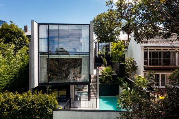 Exterior view of a contemporary multi-storey urban residence with a tall, box-like glazed facade, dark window framing, concrete or rendered side walls, and a narrow turquoise swimming pool in the landscaped courtyard. A more traditional neighbouring dwelling is visible on the right.