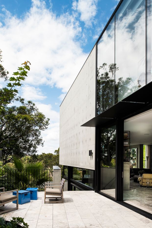 Exterior architectural view of a contemporary multi-level home with a minimalist pale concrete wall, extensive floor-to-ceiling glazing, dark metal frames, and a projecting black canopy. A paved terrace with outdoor seating connects directly to the interior living area, with landscaped greenery and a railing visible beyond.