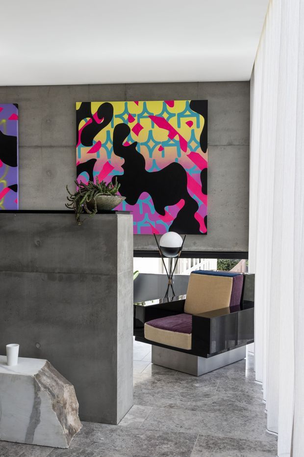 A dramatic contemporary interior with exposed grey concrete walls, a polished concrete floor, and a bold abstract artwork in vivid pink, yellow, turquoise, purple, and black. A sculptural black lounge chair with tan and plum upholstery sits beyond a partial concrete partition, accompanied by a spherical lamp and potted plant. White sheer curtains frame the right side, while daylight enters through the opening behind the seating area.
