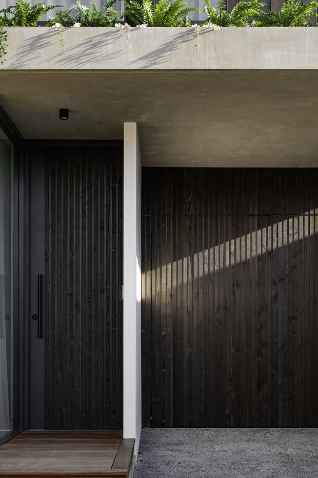 A close-up exterior view of a contemporary residential entry and garage frontage. The facade combines dark vertically slatted timber cladding, a recessed dark entry door, a pale concrete overhang and column, and a textured concrete planter above with lush ferns. Strong sunlight creates diagonal shadows across the timber garage door and wall.