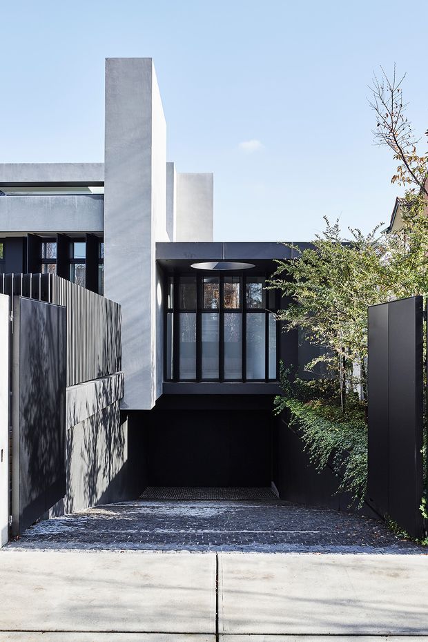 Street-facing view of a contemporary multi-level residence with a strong geometric facade. The building combines dark charcoal cladding and metal fencing with tall pale concrete-rendered vertical elements, extensive black-framed glazing, a recessed garage or undercroft, and a textured dark driveway. Sparse landscaping screens the right side, while a circular ceiling light is visible beneath the projecting upper volume.