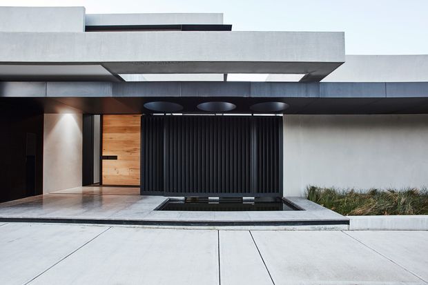 Contemporary minimalist residential facade with layered cantilevered concrete forms, a dark vertical slatted entry screen, warm timber wall accents, recessed downlights, and a shallow reflecting pool set within a pale paved forecourt. Low ornamental planting borders the right side of the entrance.