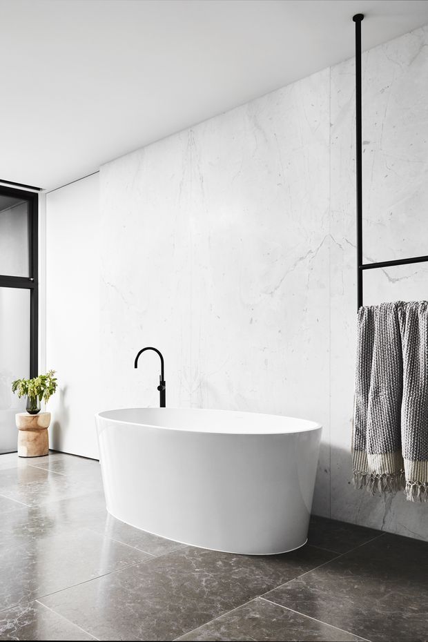 A minimalist modern bathroom featuring a freestanding white oval bathtub, black floor-mounted curved tap, large white marble-look wall panels, dark grey marble-look floor tiles, and a black-framed window. A black towel rail with woven grey towels and a potted plant add subtle contrast.