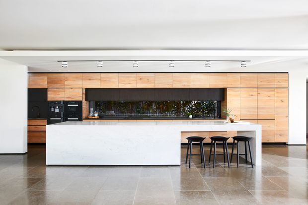 A spacious contemporary kitchen dominated by a long timber-lined wall of cabinetry and a large white stone island with integrated seating. Black appliances and dark open shelving create contrast, while polished large-format flooring and a linear ceiling track light reinforce the clean, minimalist composition.