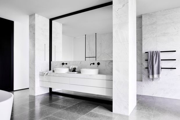 A spacious contemporary bathroom featuring a long floating white double vanity with two integrated basins, large-format pale marble-look wall surfaces, dark grey stone-look floor tiles, black-framed glazing or trim, and minimalist black wall-mounted tapware and towel rails. A freestanding white bathtub is partially visible at the left edge. The room has a clean, monochromatic and refined appearance.