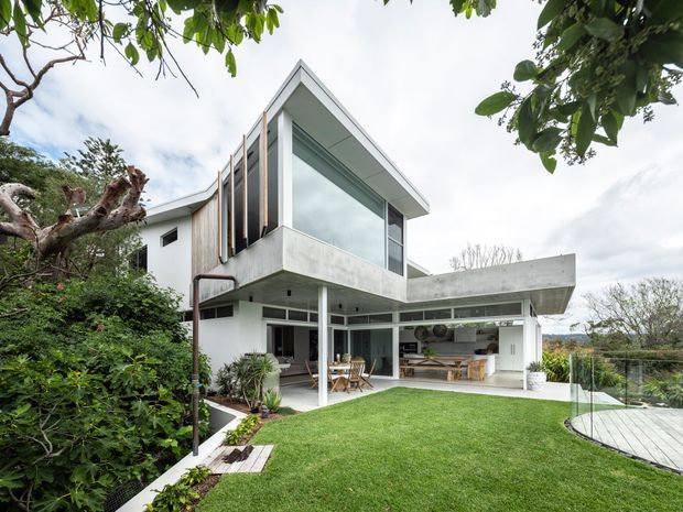 Exterior view of a contemporary multi-level residence with strong cantilevered concrete forms, extensive floor-to-ceiling glazing, and an open ground-floor entertaining area. The house opens onto a manicured lawn and timber deck, with mature vegetation framing the site. Visible interiors include an outdoor-facing kitchen or serving area and a dining space.