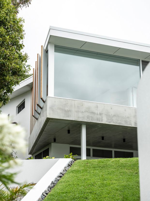 Exterior view of a contemporary multi-level home on a sloped landscaped site. The building features a flat roof, large glazed openings, white rendered walls, exposed concrete balcony and soffit surfaces, slim white structural columns, and vertical timber screening elements. A grassed incline and stone-lined pathway lead toward the partially recessed lower level.