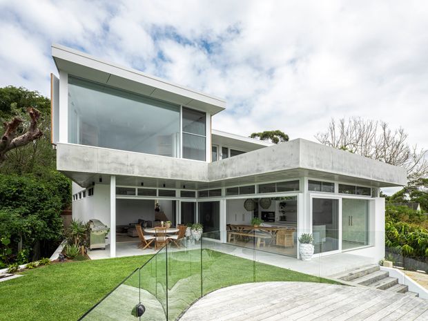 Exterior view of a contemporary two-storey residence with strong horizontal concrete forms, extensive floor-to-ceiling glazing, white structural elements, and an open-plan living area visible through the windows. The house opens onto a landscaped lawn and pale timber deck with frameless glass balustrades and outdoor steps.