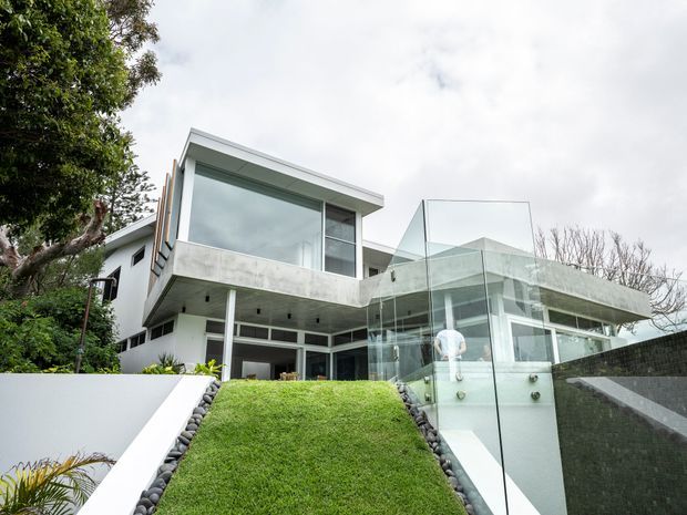 A contemporary multi-level residential house viewed from the front, featuring crisp white and concrete cantilevered forms, extensive floor-to-ceiling glazing, and a transparent glass balustrade along the upper terrace and stair access. The landscaped approach includes a steep manicured grass bank, dark stone edging, and a dark masonry retaining wall, with mature trees framing the property.