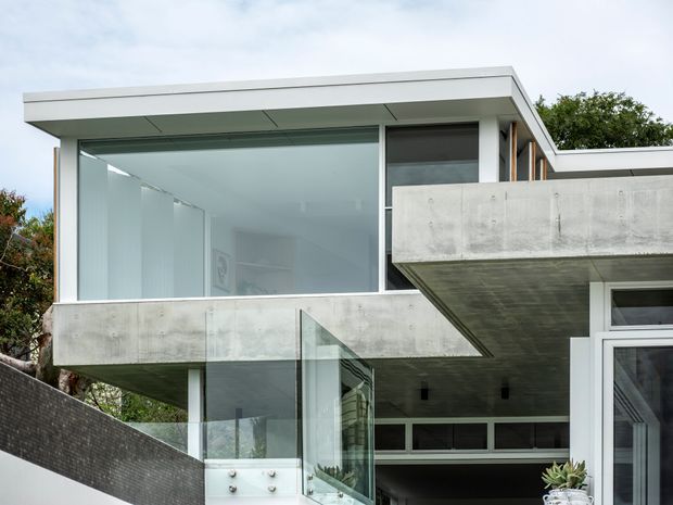 Exterior view of a contemporary multi-level residence with strong horizontal lines, broad floor-to-ceiling glazing, exposed concrete cantilevered balconies and roof slabs, and white-framed openings. A glass balustrade and partially visible outdoor pool or water feature appear at the lower level, with landscaping around the building.