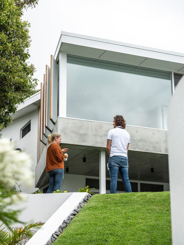 Exterior view of a contemporary multi-level residential building, featuring a crisp white flat roof, expansive glazing, exposed concrete balcony and soffit surfaces, and vertical timber screening. Two people stand on a landscaped sloping lawn in the foreground.