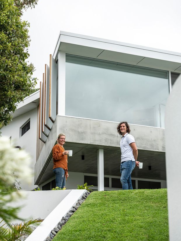 Exterior view of a contemporary multi-level residence on a sloped landscaped site. The building features crisp white geometric volumes, a flat roof, extensive floor-to-ceiling glazing, exposed raw concrete balcony and soffit surfaces, and vertical timber screening elements. Two people stand on the lawn in front of the house.
