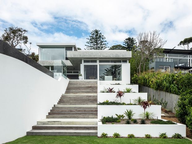 Exterior view of a contemporary multi-level residence approached by a broad timber-toned stairway. The home features crisp white rendered walls, extensive glazing, a projecting upper volume with a flat roof, and a grey concrete or masonry balcony/parapet. Terraced white planter walls with structured landscaping line the stairs, while neighbouring modern homes and mature trees are visible in the background.