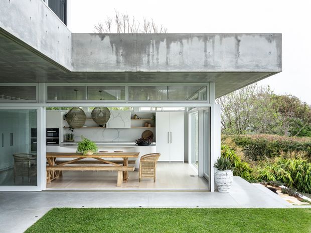 A contemporary concrete house with a cantilevered upper volume and large glazed openings, viewed from a landscaped lawn. The open ground-floor kitchen and dining area features white cabinetry, a pale stone-look splashback, timber dining table and bench seating, woven pendant lights, and direct access to an outdoor terrace and garden.