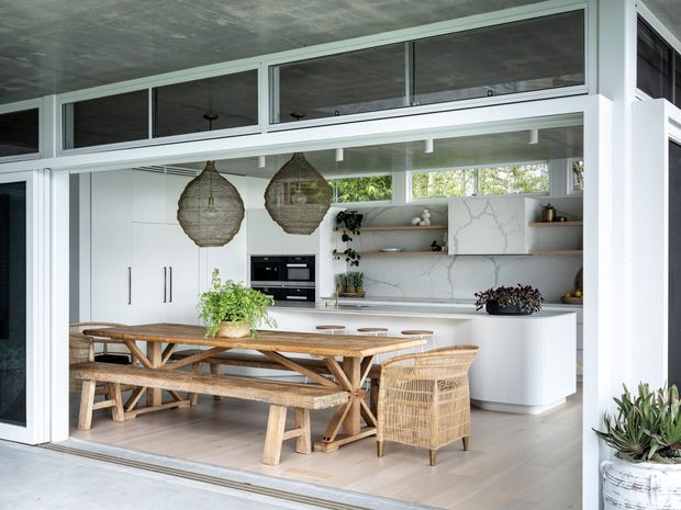 Bright open-plan kitchen and dining area viewed through fully open glazed doors. The space features white built-in cabinetry, a pale veined stone benchtop and splashback, open timber shelves, integrated appliances, a long rustic timber dining table with matching benches, a woven rattan dining chair, and two large woven pendant lights. High clerestory glazing and a textured concrete ceiling add architectural character, while indoor plants contribute a natural accent.