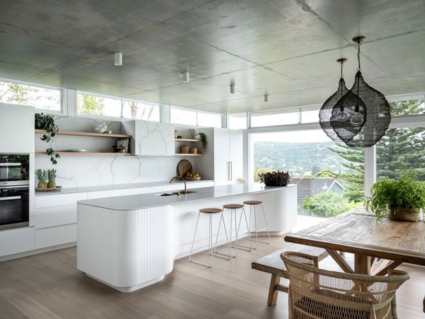 Bright contemporary open-plan kitchen and dining area with a large curved island, white integrated cabinetry, pale timber flooring, and expansive corner glazing overlooking a leafy residential landscape. The kitchen features a light stone-look benchtop and splashback, open timber shelving, minimalist appliances, and sculptural woven pendant lights. A timber dining table with woven chairs occupies the foreground.