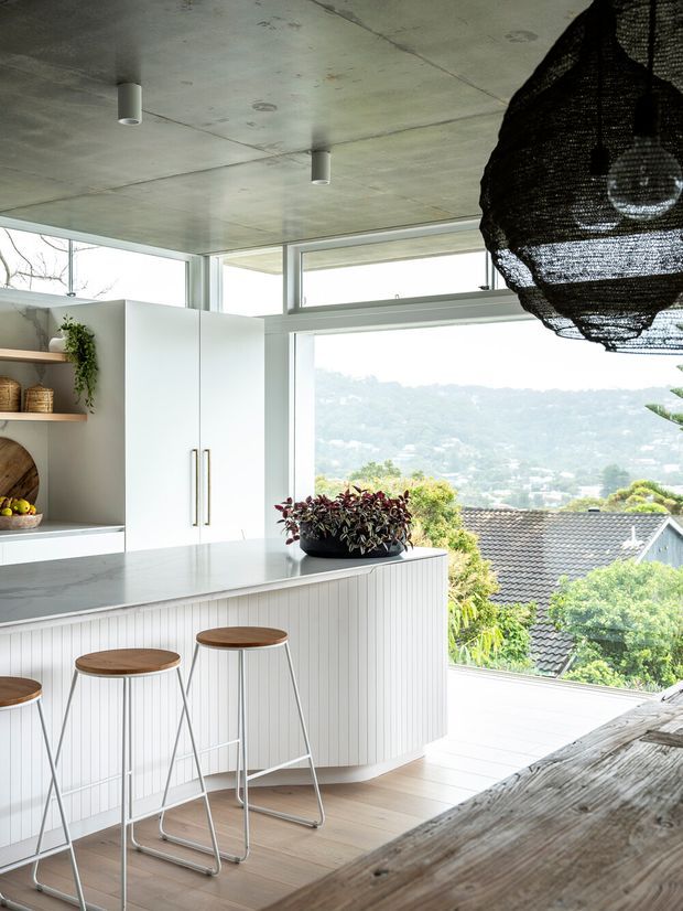 Bright contemporary kitchen with a large island clad in vertically grooved white panelling, pale stone benchtop, white full-height cabinetry, open timber shelving, and three slim timber-seated stools. Floor-to-ceiling and clerestory glazing provide expansive views over a leafy suburban hillside. A raw concrete ceiling, minimal cylindrical downlights, and a large black woven pendant add contrast, while a timber dining surface occupies the foreground.