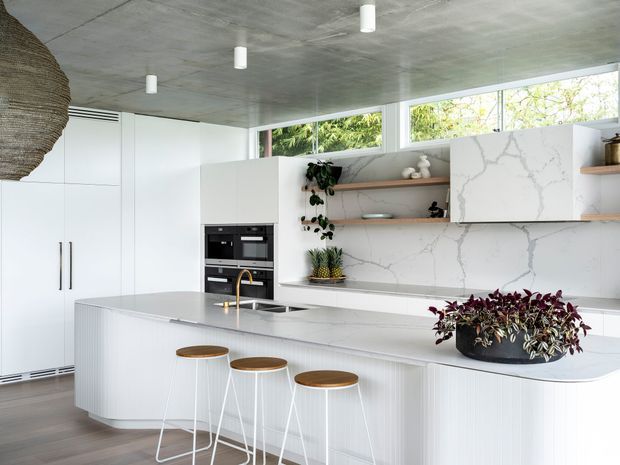 Bright contemporary kitchen with a large curved island and three stools, white integrated cabinetry, built-in black appliances, pale stone benchtops and splashback with grey veining, open timber shelves, and a raw concrete ceiling. Clerestory glazing brings in natural light, while indoor plants and a woven pendant add warmth.