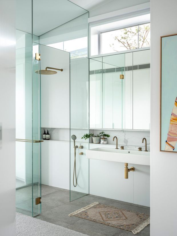 Bright contemporary bathroom featuring a frameless glass walk-in shower with brass hardware and rain showerhead, a large mirrored wall, a floating white double vanity with brass taps and exposed plumbing, and large grey floor tiles. High clerestory glazing brings in natural light, while potted plants and framed artwork add restrained decorative detail.