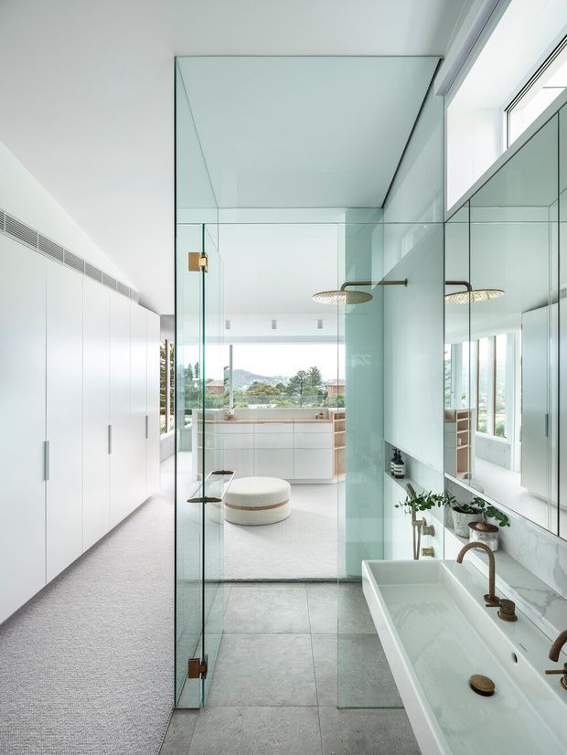 A bright, refined bathroom with a long freestanding rectangular bathtub and double vanity positioned beside a large mirrored wall. A frameless glass shower enclosure separates the bathroom from a carpeted dressing or bedroom area beyond. White full-height cabinetry lines the left wall, while pale stone-look surfaces, large grey floor tiles, brass fittings, and abundant glazing create an airy contemporary interior with views toward a leafy landscape.
