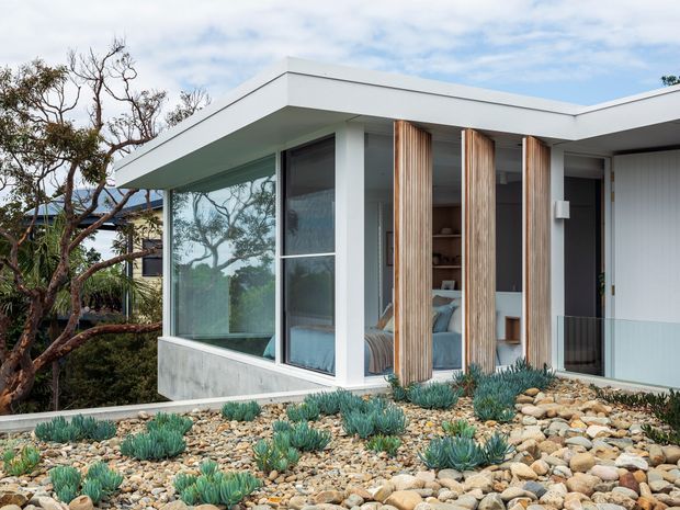 Exterior view of a low, flat-roofed modern home with extensive floor-to-ceiling glazing, white rendered walls, vertical timber screening fins, and a concrete base. The building sits among drought-tolerant succulent planting and decorative stone landscaping, with a bedroom or living area visible through the glazing.