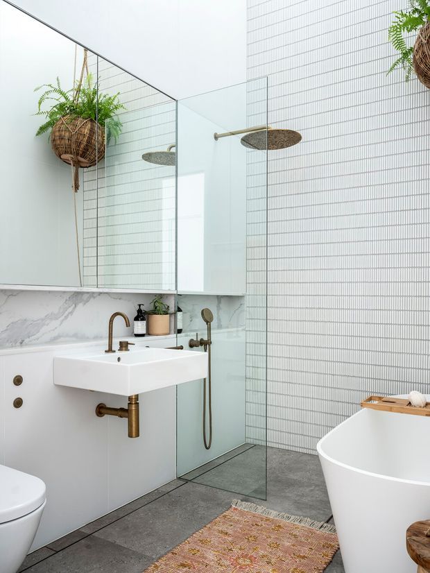 Bright contemporary bathroom featuring a wall-mounted white basin with brushed brass tapware and exposed brass plumbing, a freestanding white bathtub, glass shower enclosure, full-height mirrored cabinetry, and vertically gridded pale wall tiles. A marble-look splashback, dark grey floor tiles, woven pendant planters, and a patterned rug add texture and warmth.