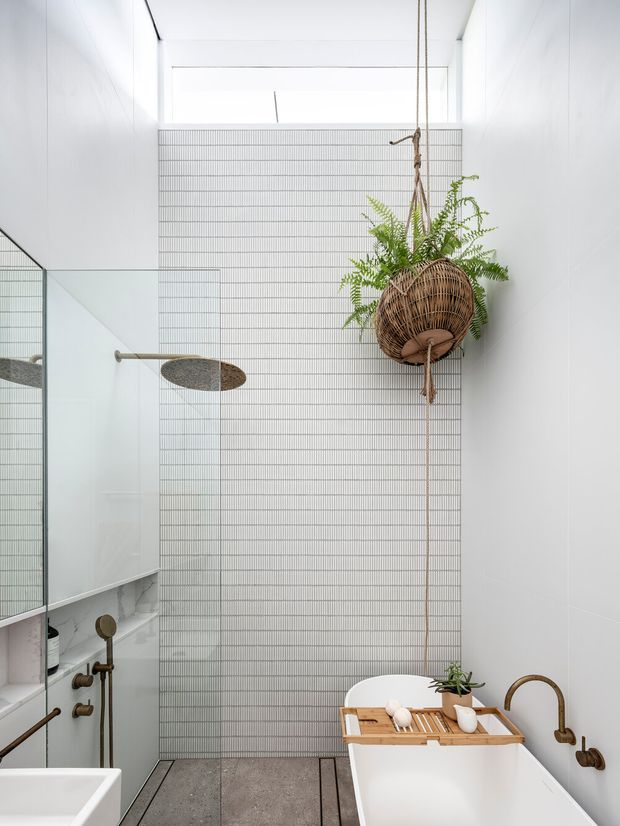 A bright, narrow contemporary bathroom with a freestanding white bathtub in the foreground, a glass-enclosed shower at left, and a floor-to-ceiling tiled feature wall made of fine horizontal lines. White walls and ceiling create an airy, light-filled space. A woven hanging planter with fern greenery adds a natural accent, while brushed brass or bronze-toned tapware and fixtures provide warmth. Large-format dark grey floor tiles contrast with the white surfaces.