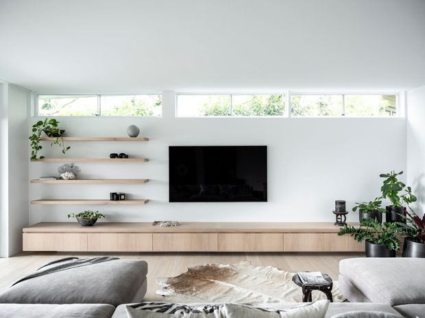 Bright contemporary living room with a low built-in timber media console, wall-mounted television, and open floating display shelves. High horizontal clerestory windows bring in abundant daylight, while grey upholstered seating, a natural hide rug, and potted plants create a calm, warm interior.