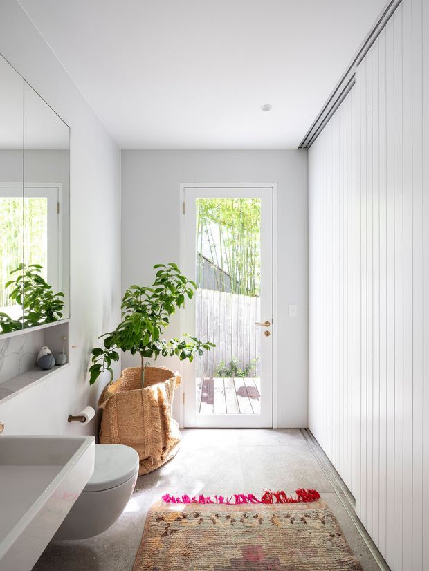 A bright, narrow contemporary bathroom with a wall-mounted white vanity and toilet, large mirror, pale tiled floor, and full-height white vertical wall panelling. A glazed exterior door opens to a small timber deck and leafy garden, bringing in abundant natural light. A woven basket, indoor plant, and colourful patterned rug add warm organic accents to the otherwise minimalist space.