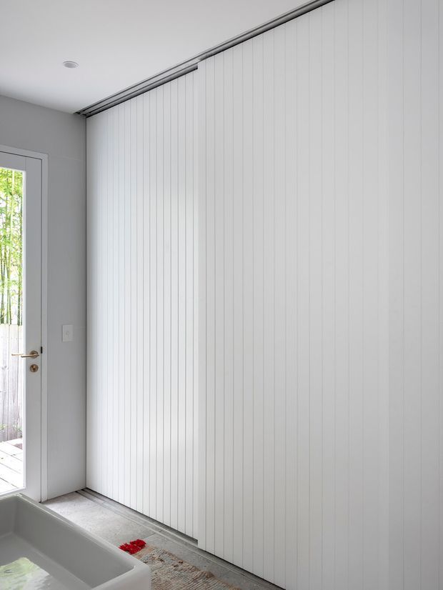 Bright minimalist interior showing a large wall of white vertically grooved sliding or folding panels, likely concealing storage or a room opening. A glazed exterior door is visible at left, with a partial white freestanding bathtub in the foreground and pale tiled flooring. The space has clean contemporary detailing and abundant natural light.