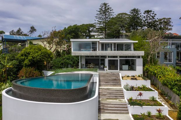 Exterior view of a contemporary multi-level house set in a lush landscaped garden. The white and grey facade has broad horizontal forms, extensive glazing, flat rooflines, and open balconies or terraces. A curved raised swimming pool dominates the foreground, alongside stepped timber-look paving and tiered white garden planters.