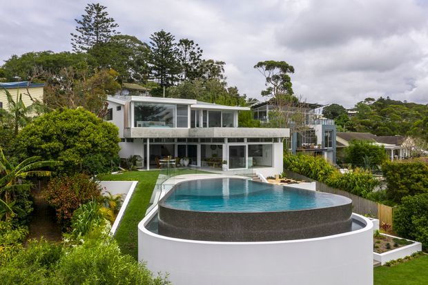 Exterior architectural photograph of a modern multi-level residence set among dense landscaped greenery. The white and grey house has extensive floor-to-ceiling glazing, projecting balconies or terraces, and a distinctive elevated curved swimming pool in the foreground with a dark rendered outer wall and white circular base. The scene has an overcast sky and lush suburban vegetation.