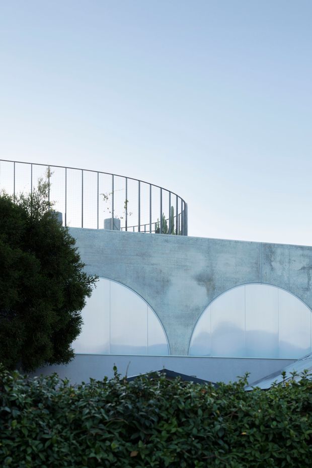 Exterior architectural photograph of a pale blue-grey curved concrete or plaster building facade, featuring large translucent arched panels and a rooftop enclosure with curved metal mesh fencing. Dense hedging and trees partially obscure the lower facade, with a clear blue sky above.