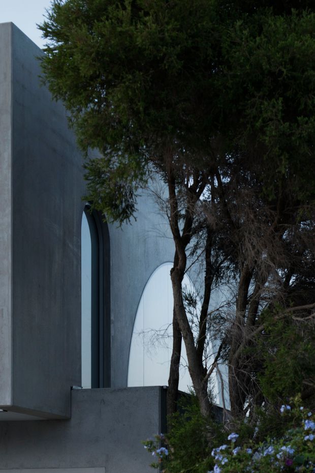 A moody exterior architectural view of a minimalist concrete building partially screened by mature trees and shrubs. The facade has smooth dark grey rendered or concrete surfaces, tall narrow arched window openings, and a prominent large curved translucent or glazed opening behind a low concrete wall. The composition emphasises the contrast between sculptural geometric architecture and dense landscaping.