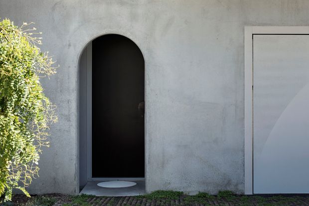 Minimal exterior facade detail featuring a smooth grey rendered wall, a tall rounded-arch dark doorway, and a large simple rectangular garage door. Low groundcover planting and subtle paving line the base, creating a restrained contemporary architectural composition.