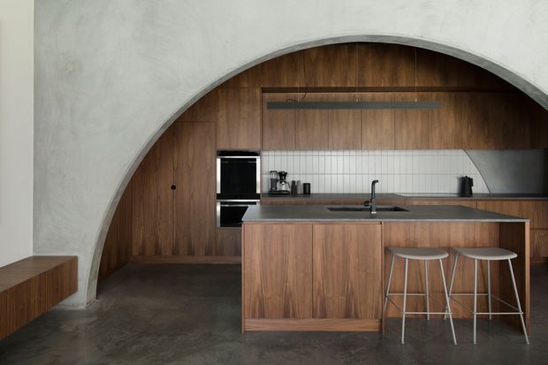 A contemporary kitchen framed by a large curved plaster arch, with warm vertical timber wall panelling and matching island cabinetry. The kitchen includes a dark stone or composite benchtop, white vertical tile splashback, integrated black appliances, a slim linear pendant light, and two metal bar stools. The polished concrete floor and restrained palette create a minimalist, refined atmosphere.