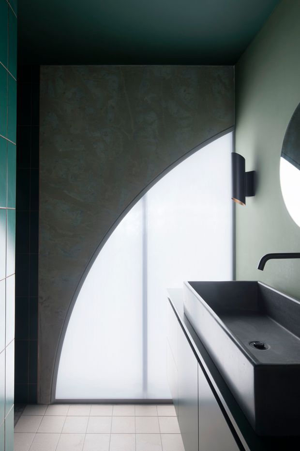 A moody, highly contemporary bathroom featuring a dark rectangular basin with a black wall-mounted tap, a large round mirror, minimalist wall lighting, and a dramatic curved architectural partition framing a tall frosted-glass opening. Dark green-black tiled walls contrast with pale floor tiles and muted green-grey surfaces.