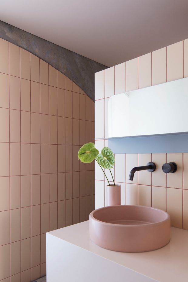 Close-up of a contemporary bathroom vanity featuring a rounded blush-pink vessel basin on a white benchtop, matte black wall-mounted tapware, and a large white curved-front mirrored cabinet or wall unit. The walls are clad in vertically arranged dusty pink rectangular tiles with fine darker grout lines, while a dark textured concrete structural beam contrasts above. A small potted plant adds a natural accent.